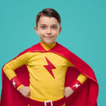 Smiling superhero child in bright cloak standing with hands on waist and looking at camera on blue background