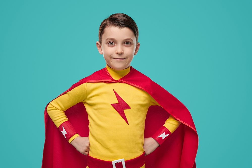 Smiling superhero child in bright cloak standing with hands on waist and looking at camera on blue background